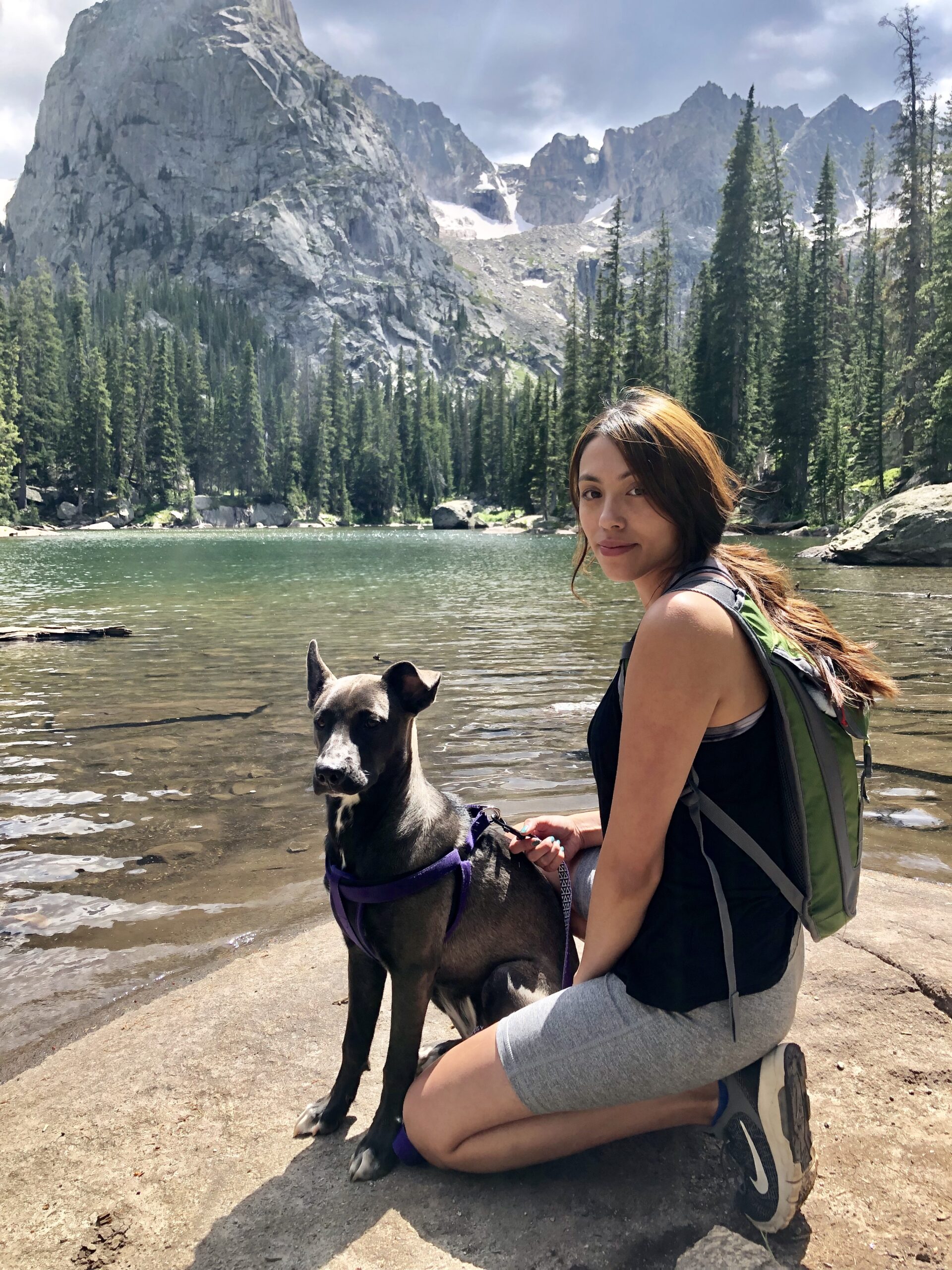 Angelica Hiking with Her Dog in the Mountains Angelica kneeling beside her dog at an alpine lake surrounded by evergreen trees and rocky mountain peaks during a summer hike.