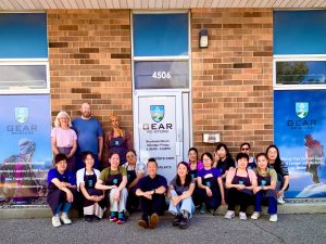 Group of Gear re-Store team members posing in front of the brick storefront at 4506, Canada's Gear re-Store's location, wearing company aprons and smiling beneath the Gear Re-Store signs.