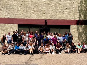 Group of about 40 people posing for a team photo in front of a tan brick, building labeled “Unit 100,” Colorado's Gear re-Store's location, with some standing and others sitting on the pavement under bright sunlight.