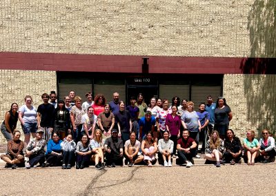 Group of about 40 people posing for a team photo in front of a tan brick, building labeled “Unit 100,” Colorado's Gear re-Store's location, with some standing and others sitting on the pavement under bright sunlight.