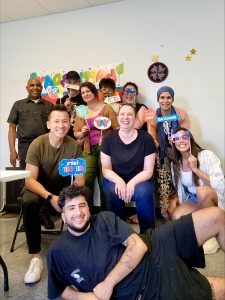 Group of ten people smiling and holding colorful party signs in front of a decorated wall with balloons, celebrating together in an office, Pennsylvania's Gear re-Store's location.