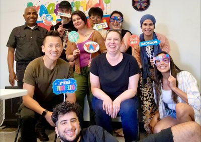 Group of ten people smiling and holding colorful party signs in front of a decorated wall with balloons, celebrating together in an office, Pennsylvania's Gear re-Store's location.