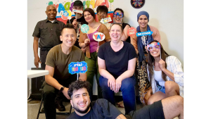 Group of ten people smiling and holding colorful party signs in front of a decorated wall with balloons, celebrating together in an office, Pennsylvania's Gear re-Store's location.